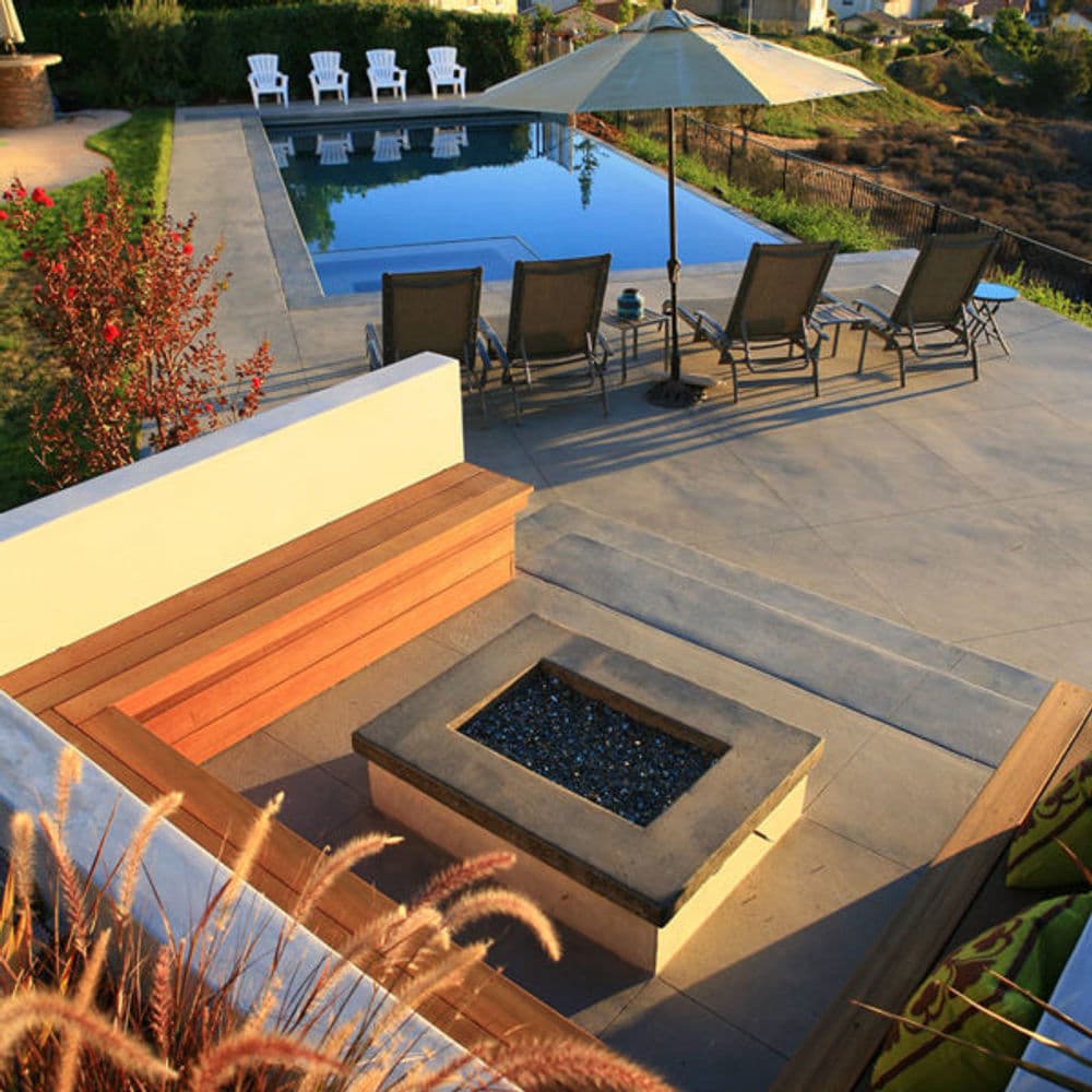 Modern outdoor patio with a pool, fire pit, lounge chairs, and umbrella in a scenic setting.