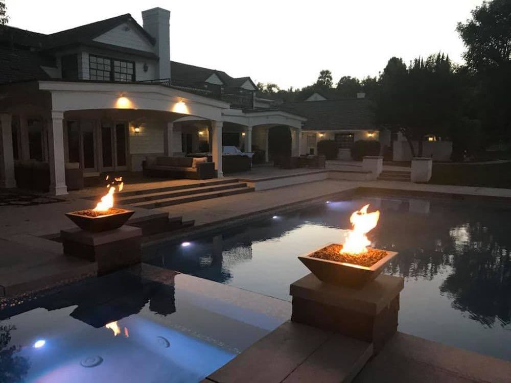 Luxury home with illuminated fire pits by a serene pool at dusk. Elegant outdoor living space.